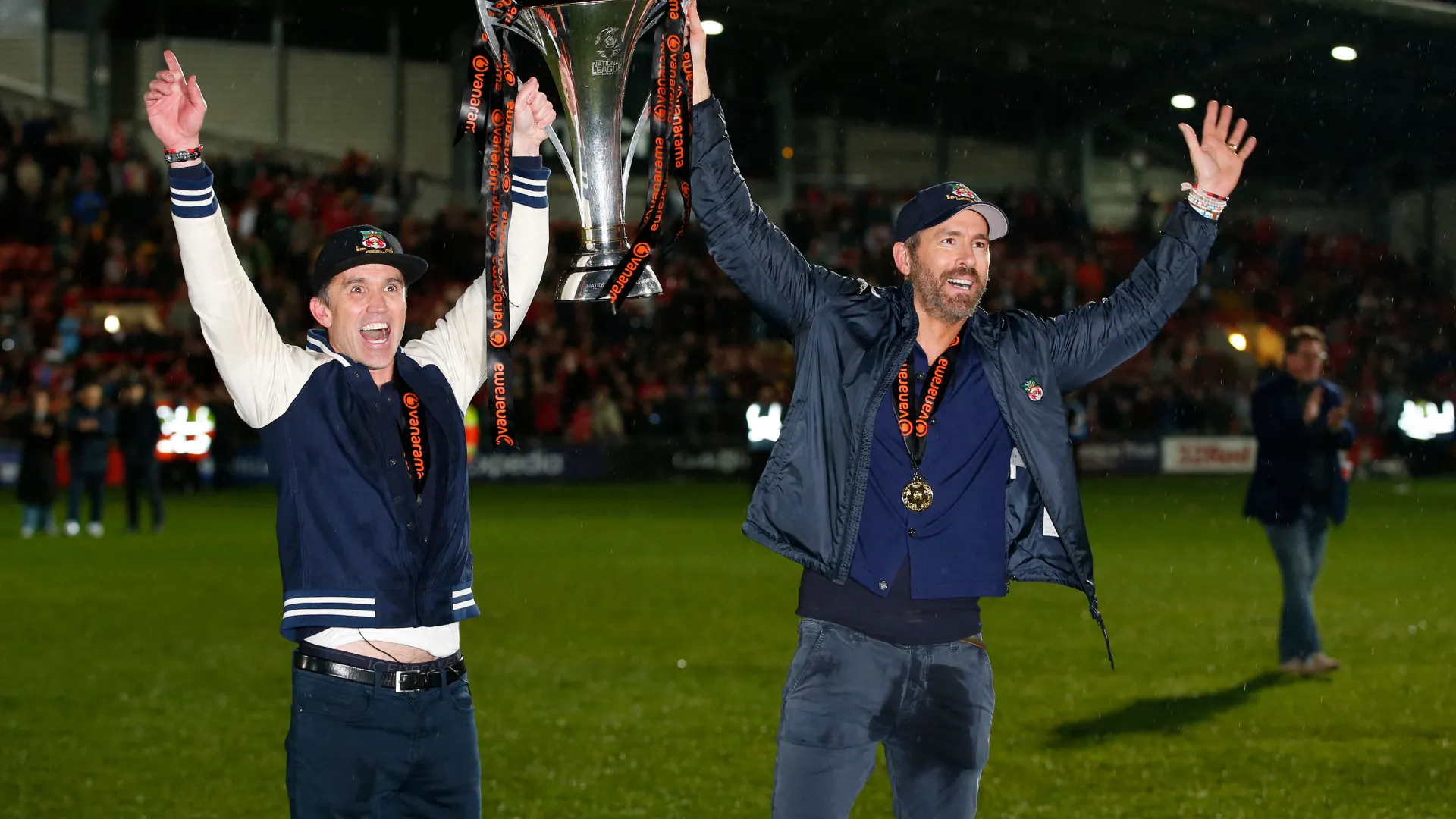 Ryan Reynolds và Rob McElhenney, hai đồng sở hữu CLB Wrexham, đứng cạnh nhau trên sân cỏ.